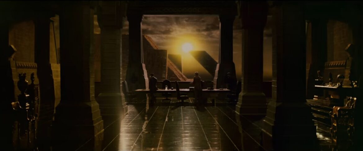 A dark cavernous office with huge pillars; beyond, the sun is visible in the dark sky.