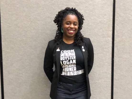 A Black woman smiles at the camera. She wears a black jacket and a tee with names of Black rhet-comp elders/scholars.