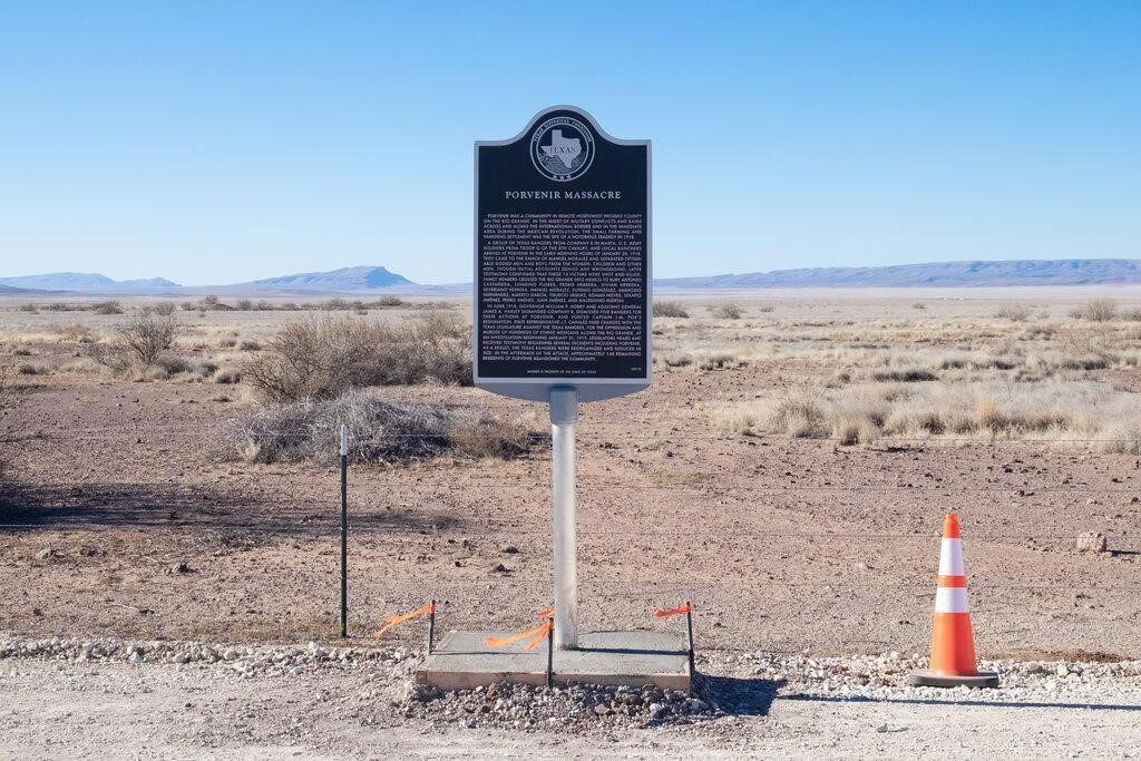 A marker accompanied by a traffic cone stands before a bared wire fence. The desert landscape leads to hills in the distance.