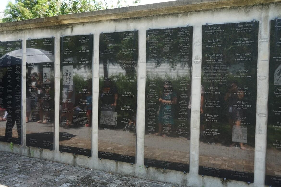 A wall holds up black marble panels recording many names and some photographs of people. The silhouettes of visitors are reflected in the marble.