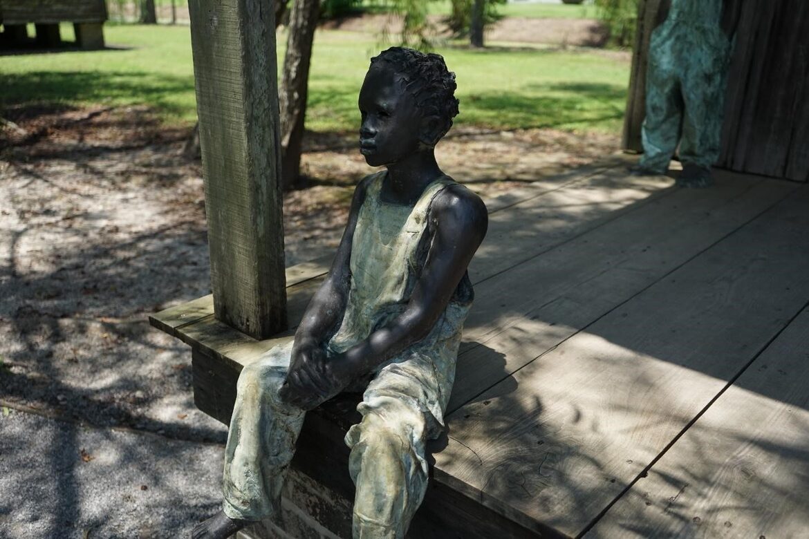 A sculpture of an enslaved Black child barefoot and dressed in simple overalls and sitting on the edge of a wooden porch.