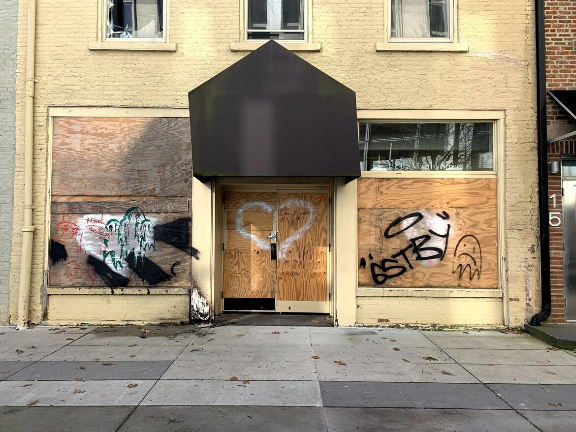 An older building with plywood-covered doors and windows. The plywood is covered in graffiti.